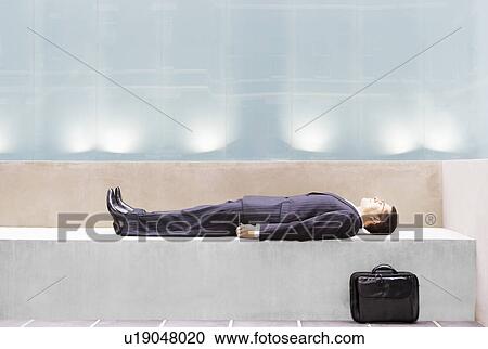 Stock Image - Businessman lying on concrete bench, side view. Fotosearch