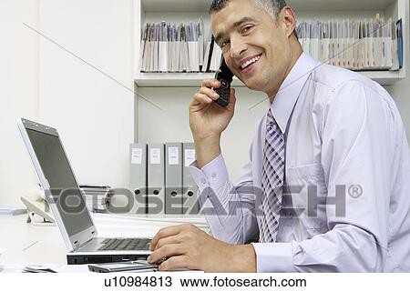 Businessman using laptop and mobile phone at desk, portrait View Large Photo Image Stock Image - Businessman using laptop and mobile phone at desk, portrait. Fotosearch