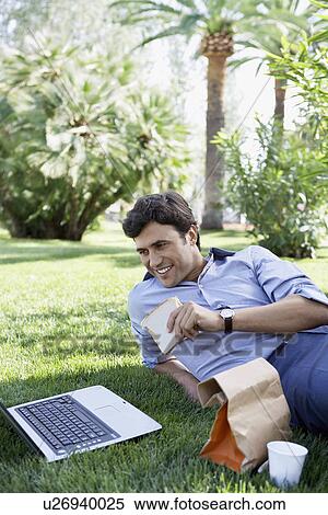 Businessman using laptop while eating sandwich in park View Large Photo Image Stock Photography - Businessman using laptop while eating sandwich in park. Fotosearch
