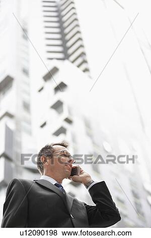 Stock Photo - Businessman using mobile phone in front of tall building, low angle view. Fotosearch