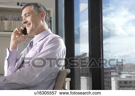 Stock Photo - Businessman using mobile phone, sitting in front of window overlooking cityscape. Fotosearch