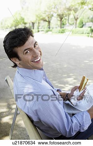 Picture - Businessman with sandwich and newspaper sitting on park bench. Fotosearch