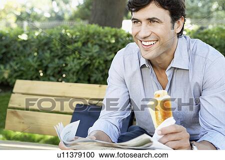 Stock Image - Businessman with sandwich and newspaper sitting on park bench, close-up. Fotosearch