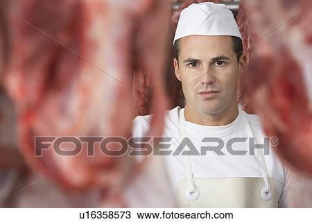 Stock Image - Butcher Standing in Meat Locker, behind raw slabs of meat. Fotosearch