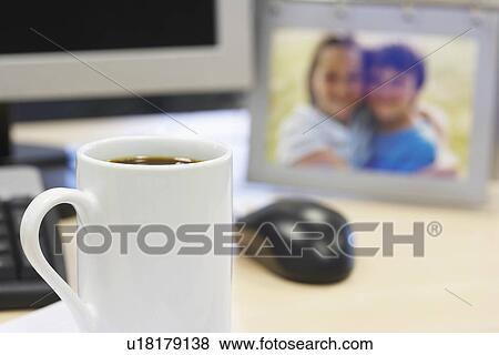 Stock Photo - Coffee mug on desk by computer and family photo, close up of mug. Fotosearch