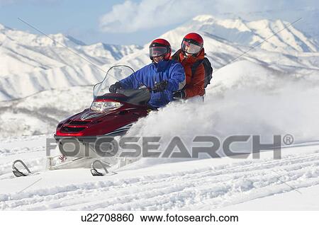 Stock Image - Couple driving snowmobile in front of snow covered mountains. Fotosearch