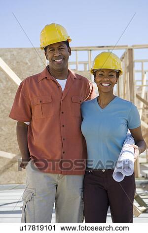 Stock Image - Couple holding building plans on construction site. Fotosearch