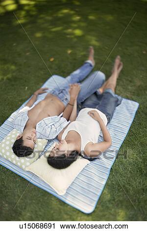 Stock Image - Couple lying down side by side on picnic blanket in park, elevated. Fotosearch