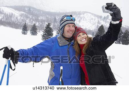 Stock Image - Couple on skis, photographing selves using digital camera, in snow covered field. Fotosearch