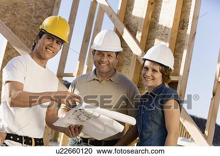 Stock Image - Couple standing with construction worker at construction site, portrait. Fotosearch