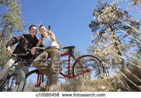 Couple standing with mountain bikes, using mobile phone, photographing selves, low angle view. View Large Photo Image Stock Photo - Couple standing with mountain bikes, using mobile phone, photographing selves, low angle view.. Fotosearch