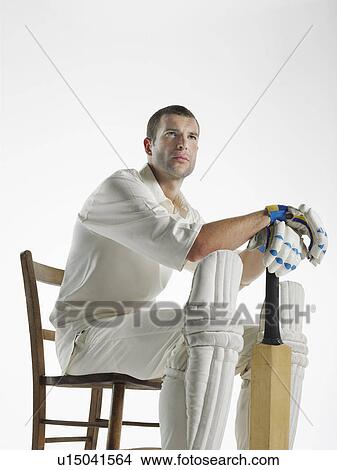 Picture - Cricket player sitting in chair, holding cricket bat, side view. Fotosearch