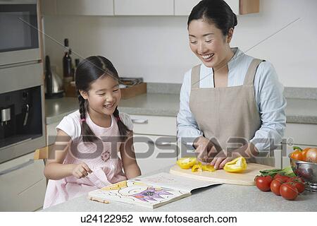 Stock Image - Daughter coloring on kitchen counter, mother cutting bell pepper and watching. Fotosearch