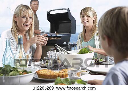 Family sitting around table outside eating Barbecue, father standing at grill grilling View Large Photo Image Stock Photo - Family sitting around table outside eating Barbecue, father standing at grill grilling. Fotosearch