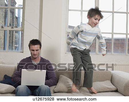 Stock Photo - Father Using Laptop as Son Jumps on Sofa. Fotosearch