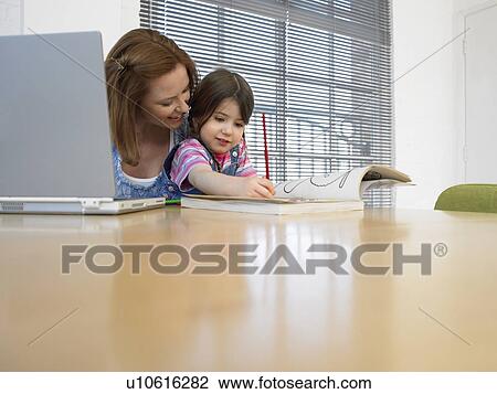 Stock Image - Girl (3-4) colouring in book beside mother using laptop, in home. Fotosearch