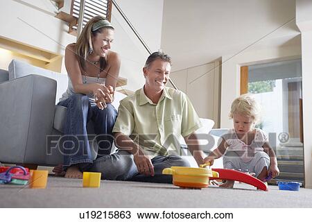 Stock Image - Happy parents watching toddler playing with toys on living room floor. Fotosearch