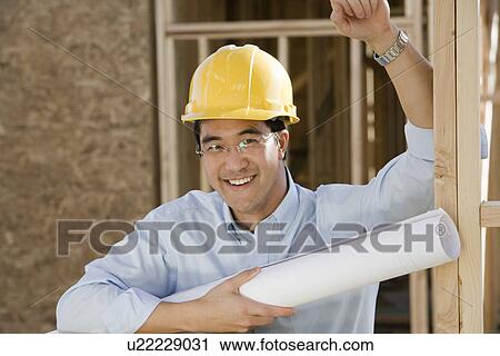Stock Image - Man holding rolled up blueprint in construction site. Fotosearch