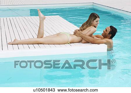 Stock Image - Man in pool hugging girlfriend on pool deck, side view. Fotosearch