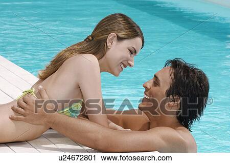 Stock Image - Man in pool hugging girlfriend on pool deck, side view. Fotosearch