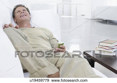 Stock Photo - Man listening to MP3 player on sofa in living room. Fotosearch
