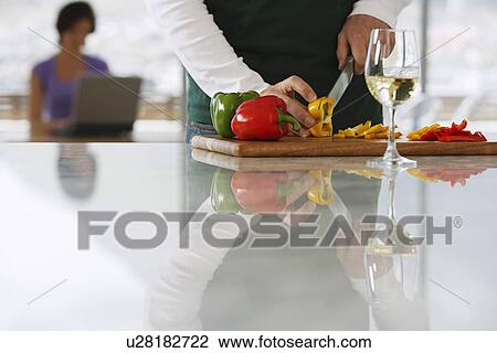 Stock Image - Man (mid section) chopping peppers in front of woman on lap top in kitchen, focus on foreground. Fotosearch
