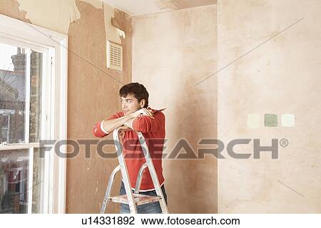 Stock Image - Man resting on step ladder in unrenovated room, looking through window. Fotosearch