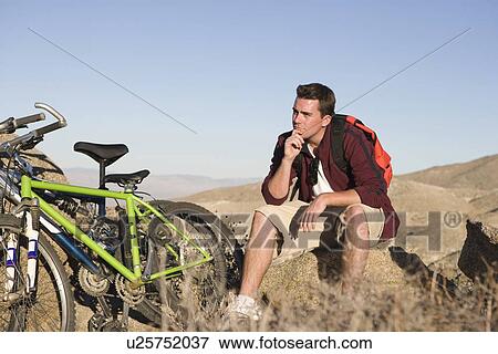 Man sits on rock thinking with mountain bikes View Large Photo Image Stock Photo - Man sits on rock thinking with mountain bikes. Fotosearch