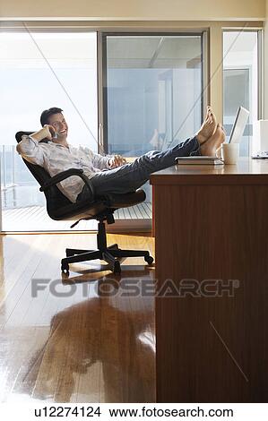Man sitting in office with feet up on desk, using mobile phone. View Large Photo Image Picture - Man sitting in office with feet up on desk, using mobile phone.. Fotosearch