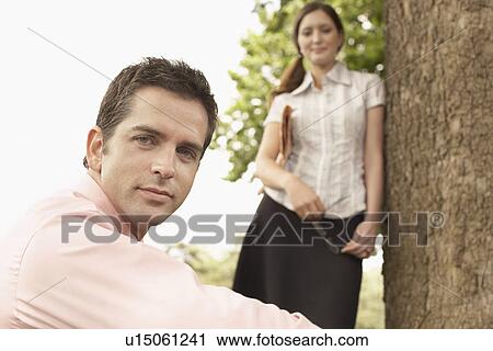 Stock Image - Man sitting in park with woman standing in background, portrait.. Fotosearch