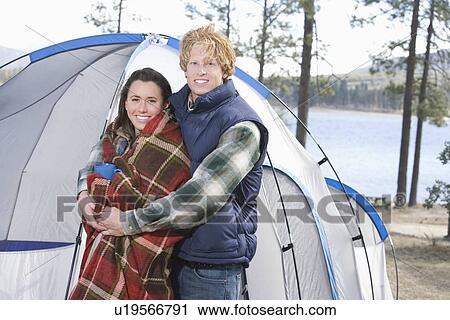 Stock Image - Man stands with arms around woman wrapped in blanket beside tent. Fotosearch