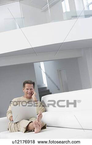 Stock Photo - Man using laptop on sofa in living room. Fotosearch