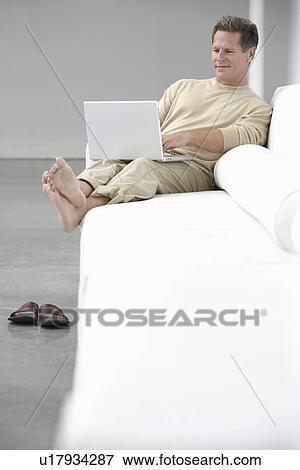 Stock Photo - Man using laptop on sofa in living room. Fotosearch
