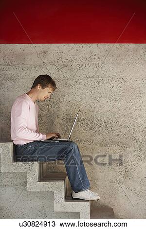 Stock Image - Man using laptop sitting on stairs, side view. Fotosearch