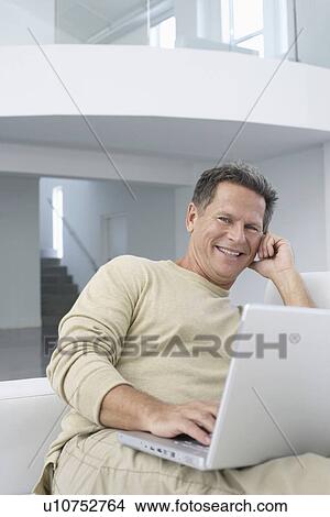 Picture - Man with laptop in living room, smiling, portrait. Fotosearch