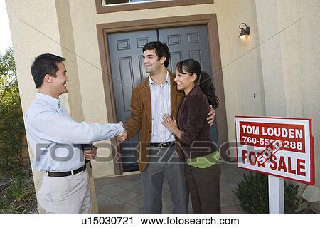 Stock Image - Mid adult couple shaking hands with estate agent in front of their new home. Fotosearch