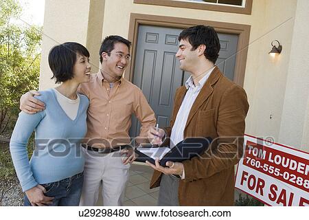 Stock Image - Mid adult couple with estate agent n front of their new home. Fotosearch