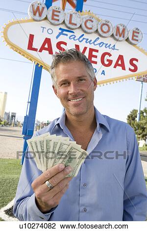 Stock Image - Mid-adult man holding notes in front of Welcome to Las Vegas sign, portrait. Fotosearch