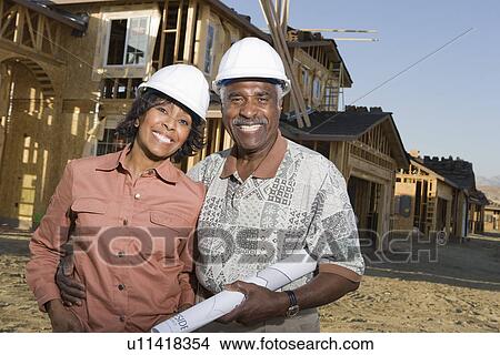 Picture - Middle-aged couple holding blueprints at construction site, portrait. Fotosearch