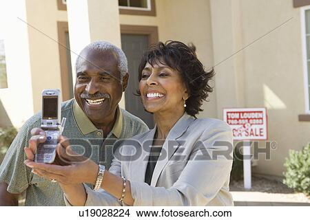 Picture - Middle-aged couple photographing in front of new home. Fotosearch