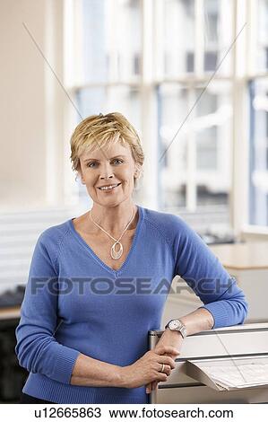 Stock Image - Middle-aged female office worker leaning on cubicle in office, portrait. Fotosearch