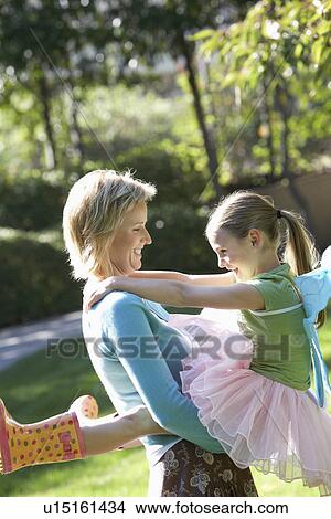 Picture - Mother holding up daughter wearing tutu in park, side view. Fotosearch