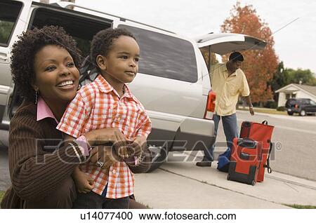 Stock Image - Mother holding young son by car, father unloading baggage. Fotosearch