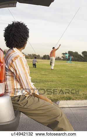 Stock Image - Mother sitting on open car boot watching father and son fly kite. Fotosearch