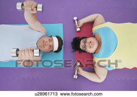 Stock Image - Overweight man and woman lying down lifting dumbbells, overhead view. Fotosearch