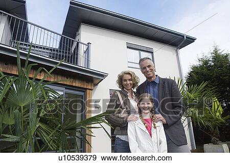 Stock Photo - Parents and daughter (7-9) embracing outside new home, portrait. Fotosearch