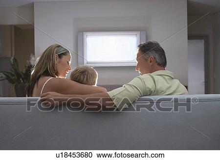 Stock Image - Parents sitting on sofa, arm around son, Watching TV, back view. Fotosearch