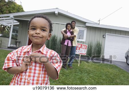 Stock Photograph - Portrait of boy (5-6) in front of new house, parents in background. Fotosearch
