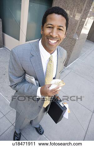 Stock Image - Portrait of businessman with newspaper and sandwich, smiling. Fotosearch