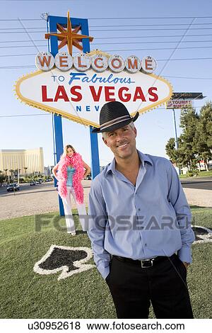 Stock Photo - Portrait of mid-adult man in front of Welcome to Las Vegas sign, mid-adult woman behind.. Fotosearch